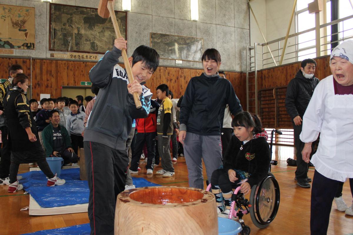 楽しそうにもちつきをする小学生とそれを笑顔で見守る先生や大熊フレンズの方