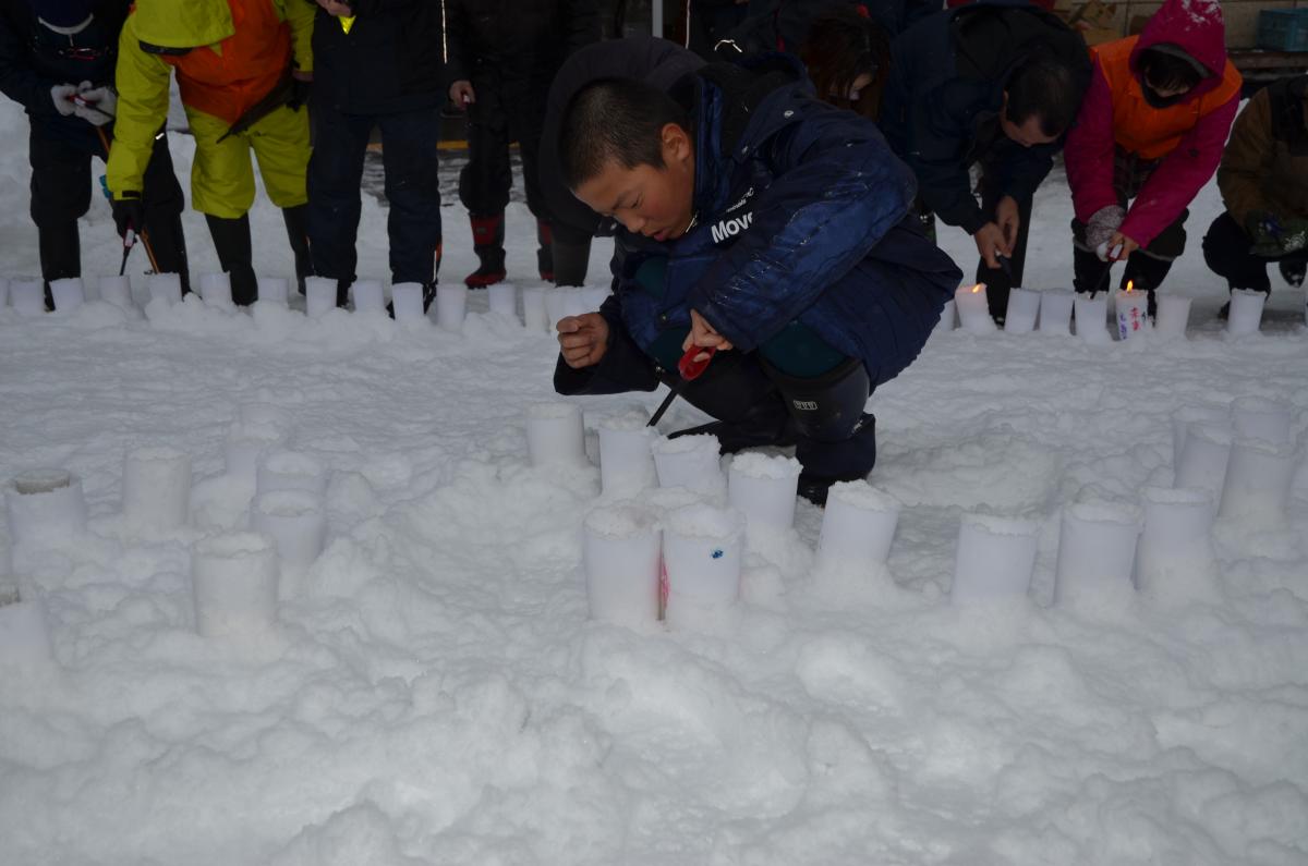 雪が積もり真っ白になった前庭に並べたろうそくに火を灯す参加者たち