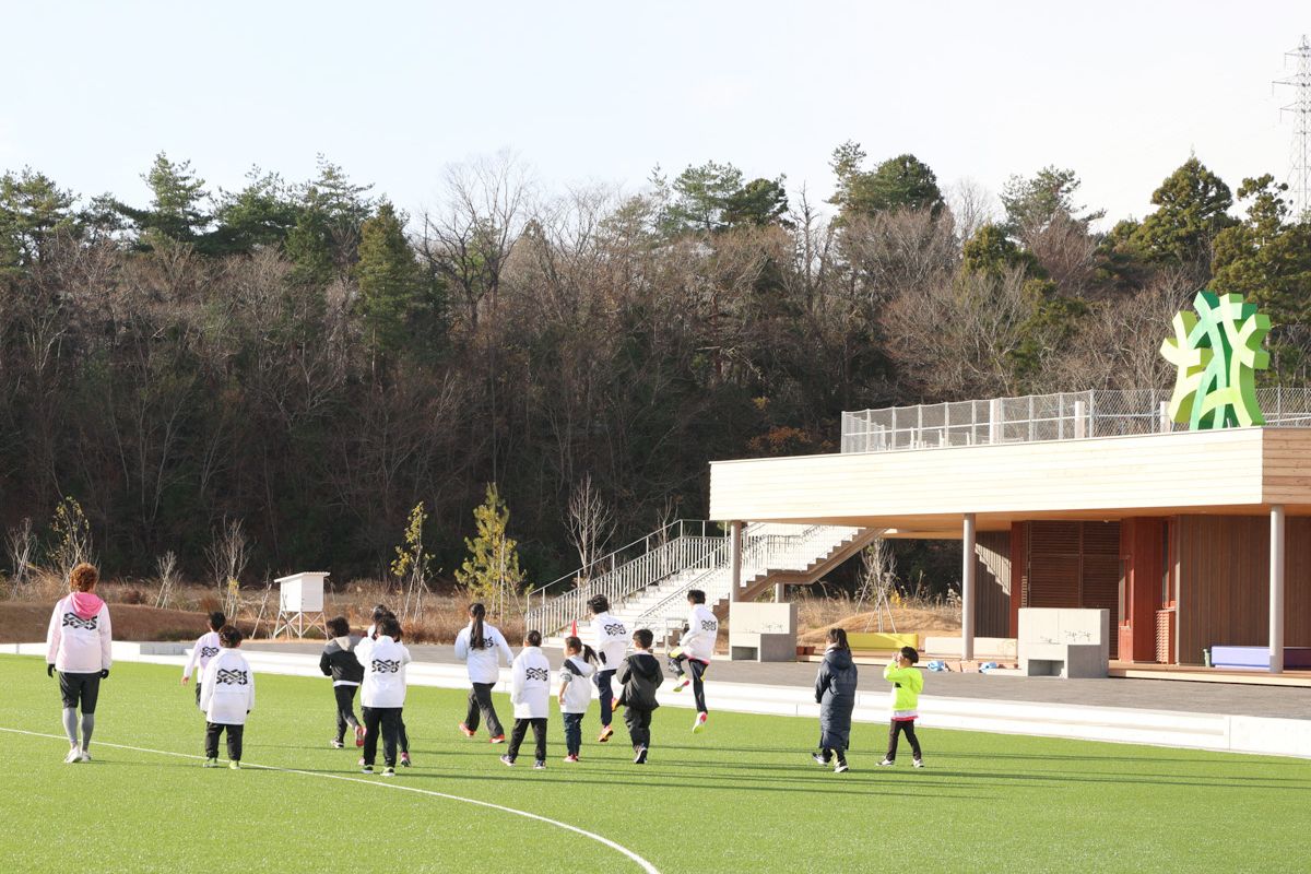 おおくま駅伝は、町立学び舎ゆめの森をスタートとゴールに、大川原地区復興拠点内の町交流ゾーンや公営住宅周辺を回るコースで行われ、町内外から出場者が集まり、小学生の部32チーム、一般の部48チーム、合わせて320人が出場。両部とも1チーム4人で、小学生の部は1.5km 、一般の部は2kmを周回してタスキをつなぎました。写真は、学び舎ゆめの森に集まった出場者が、グラウンドでウォーミングアップする様子。