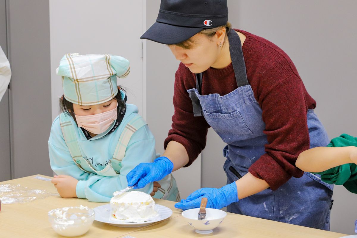 スポンジを3段重ねたらケーキの側面にもまんべんなく生クリームを塗り広げます。池田さん（写真右）は、各作業テーブルを回りながら子どもたちに塗り方のコツを丁寧に教えました。