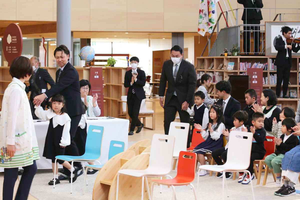 入園した3人の園児は、家族に手を引かれながらデザイナー（保育教諭）の先導で図書広場に入ると、会場いっぱいに響くあたたかい拍手で迎えられました。